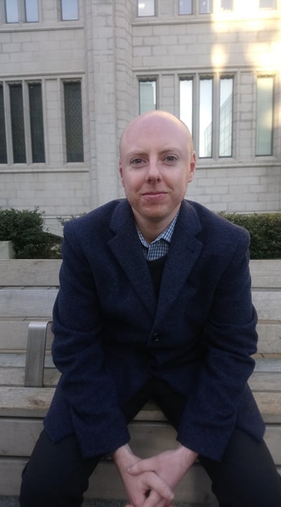 Iain Robertson is wearing dark blue jacket. He is sitting outside on the bench and looking at the camera