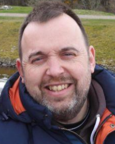 Chris Smillie has dark hair and a short trimmed beard. he is wearing a warm jacket. Behind him there is a green field
