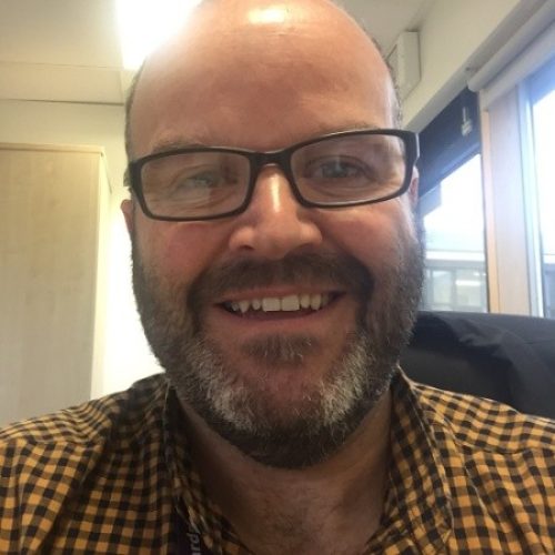 Graham is wearing glasses and yellow checked shirt. He is looking at the camera and smiling
